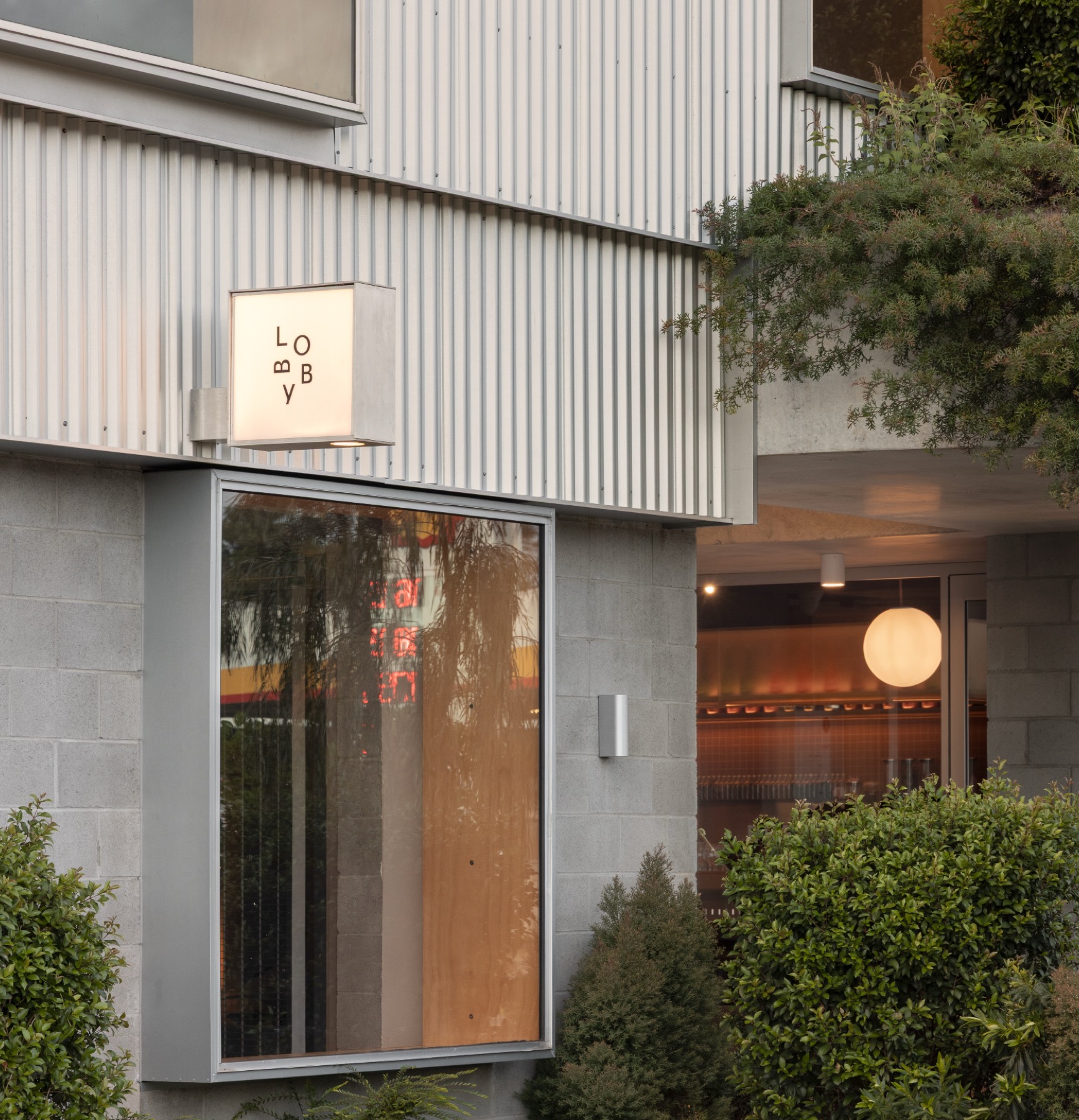 Dusk at The Warehouses, a glowing lobby lightbox on corrugated cladding beneath a planted terrace