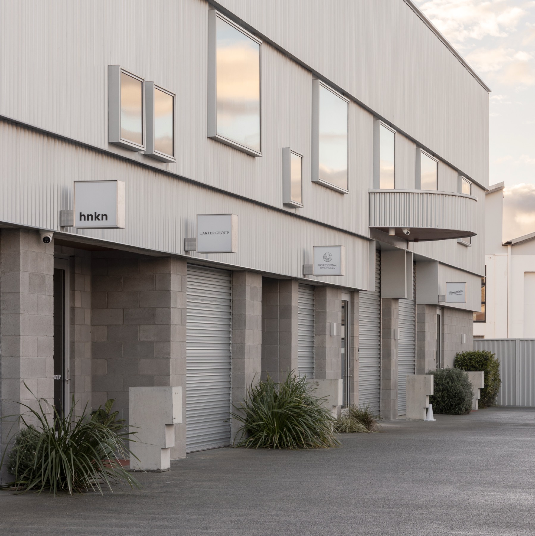 Street elevation of The Warehouses at dusk, white corrugated facade with tenant signboards and grasses against a darkening sky